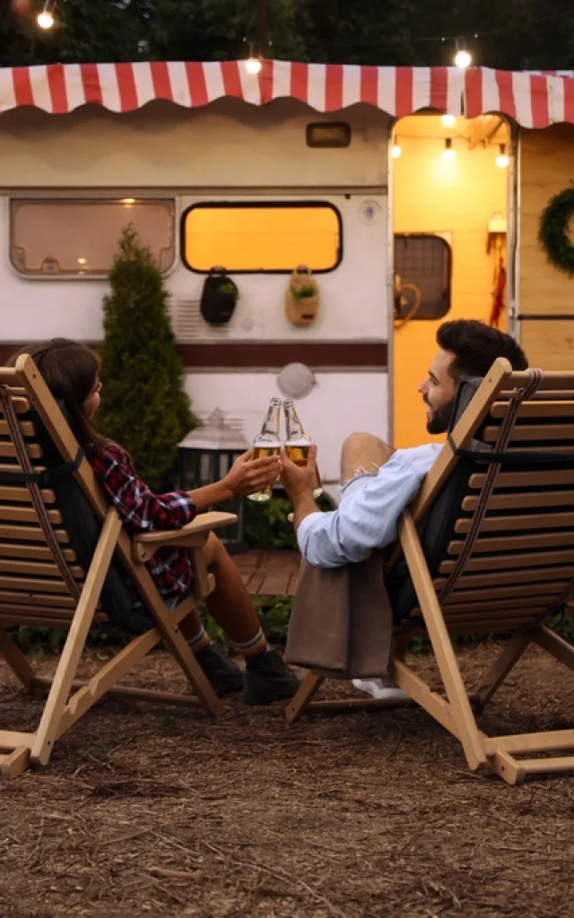 Young couple toasting with bottles of beer near trailer. Camping season